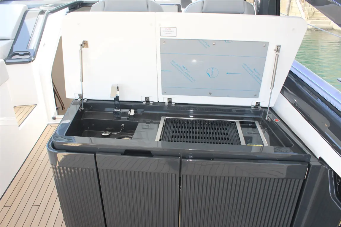 A sleek, grey boat kitchen unit with an open lid, featuring a sink, grill surface, and visible text warning.